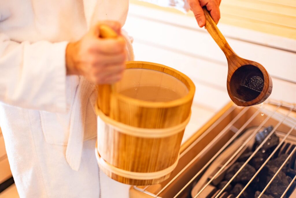 Holzeimer wird von einer Person in einem weißen Bademantel gehalten, während sie mit einem langen Holzlöffel rührt. Das Bild vermittelt eine entspannende Atmosphäre, ideal für Wellness- und Spa-Themen.