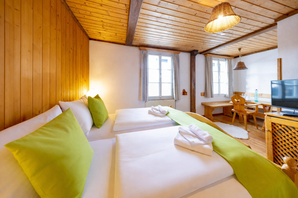Helles und modernes Gästezimmer mit zwei Einzelbetten, grünen Kissen und handtuch auf den Betten, Holzvertäfelung an den Wänden und großen Fenstern, die für viel Tageslicht sorgen. Ideal für einen entspannten Aufenthalt.
