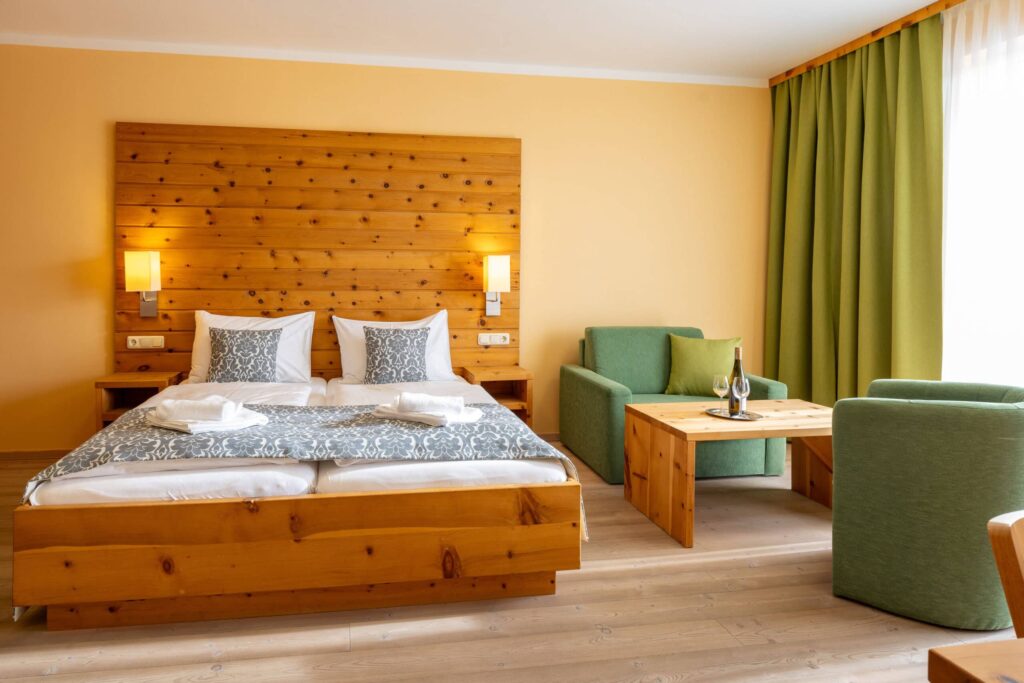 Gemütliches Hotelzimmer mit einem Doppelbett, Holzwandverkleidung und modernen Möbeln. Das Zimmer verfügt über einen Sitzbereich mit grünen Sofas und einem Holztisch, ideal für Entspannung und Komfort. Helle Wände und große Fenster sorgen für eine freundliche Atmosphäre.