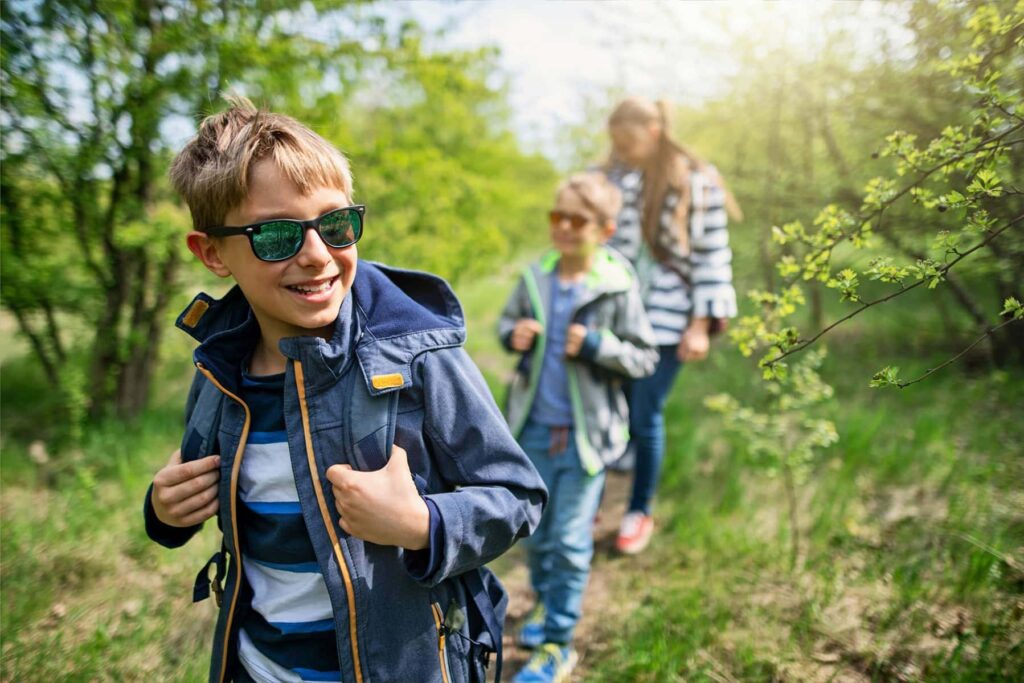 Kinder beim Wandern in der Natur, lächelnd und in wetterfester Kleidung, während sie einen grünen Waldweg erkunden. Im Hintergrund sind weitere Kinder zu sehen, die ebenfalls Ausflüge genießen.