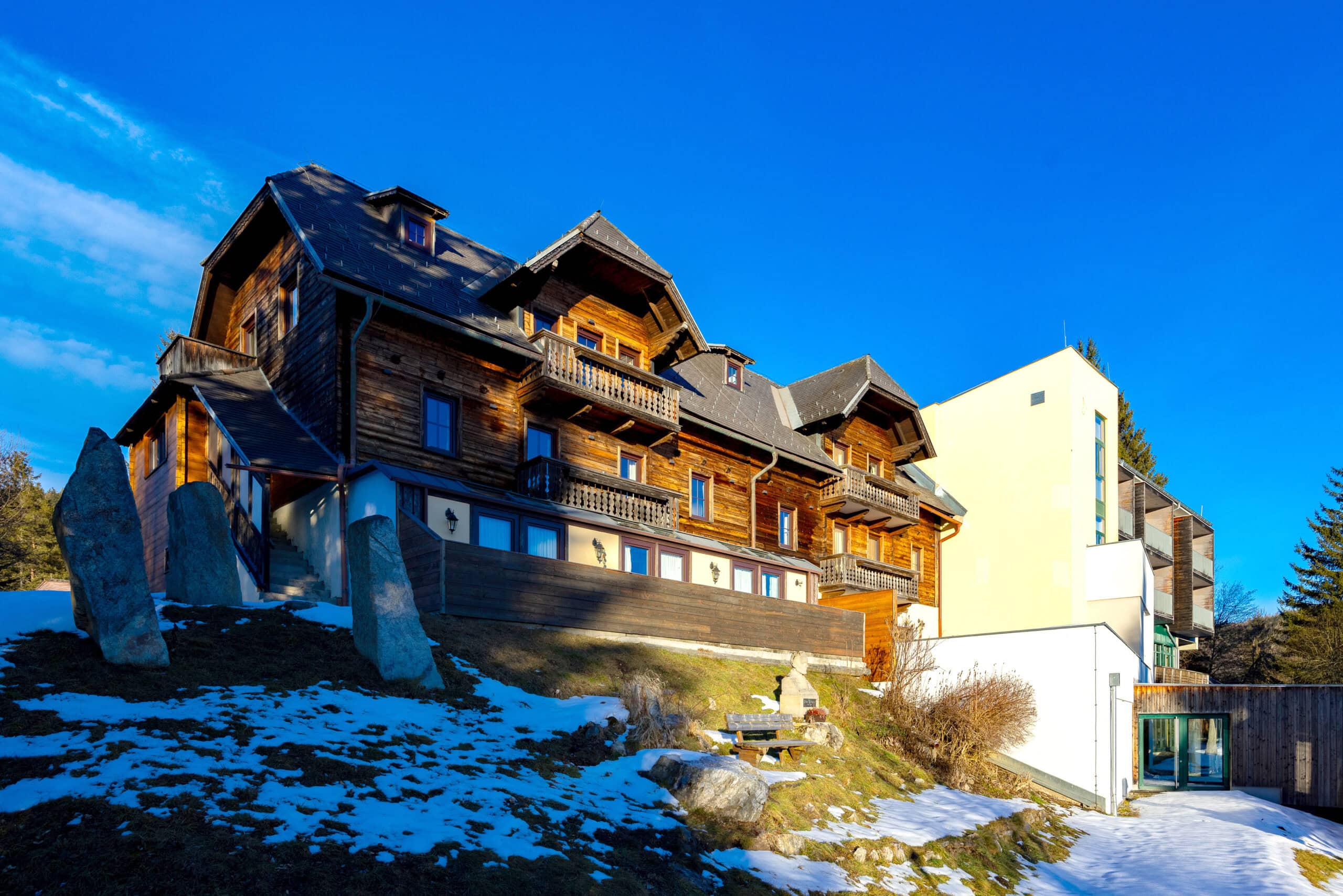 Ansicht eines modernen Holzhauses mit mehreren Etagen, das sich an einem Hang befindet, umgeben von schneebedecktem Gras und Felsen. Im Hintergrund ist ein blauer Himmel zu sehen, der die Architektur des Hauses betont. Die Fassade kombiniert traditionelle und moderne Elemente, was einen ansprechenden Kontrast schafft.