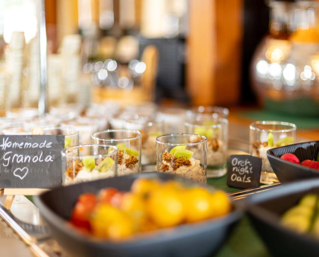 Frühstücksbuffet mit hausgemachter Granola in Gläsern, serviert mit frischen Früchten und Haferflocken. Im Vordergrund eine Schale mit bunten Beeren und Zitrusfrüchten.