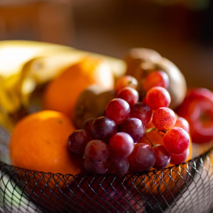 Ein Korb mit frischen Früchten, darunter Bananen, Orangen, rote Trauben, Äpfel und Kiwis, im Vordergrund unscharf dargestellt, sorgt für einen appetitlichen Anblick und vermittelt eine gesunde Ernährung.