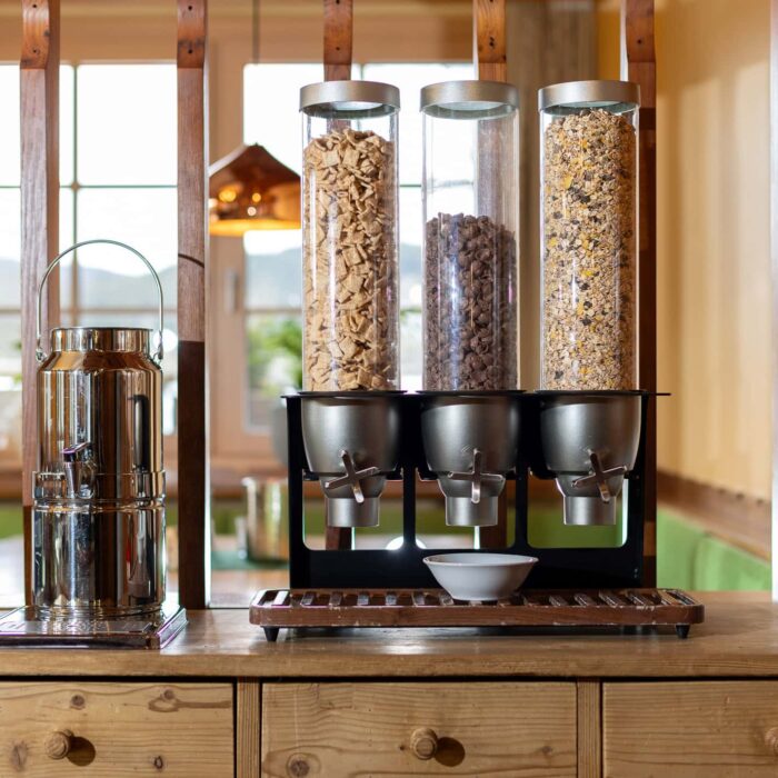 Moderne Müsli- und Snackstation mit drei transparenten Behältern für verschiedene Cerealien und Nüsse, ergänzt durch eine stilvolle Edelstahlkanne. Ideal für Frühstücksbuffets oder als praktische Aufbewahrungslösung in der Küche.