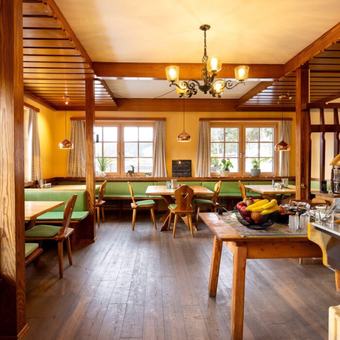 Gemütliches Restaurantinterieur mit Holzmöbeln, grünem Polster und großen Fenstern, die Tageslicht hereinlassen. Der Tisch ist mit frischem Obst und einem Frühstücksbuffet gedeckt, ideal für ein einladendes Frühstückserlebnis.