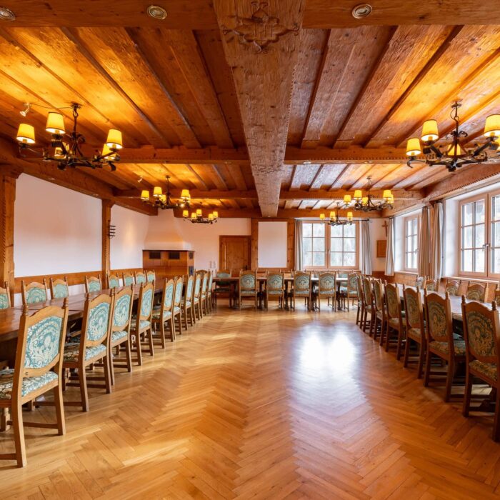 Eleganter Tagungsraum mit Holzdecke und -boden, ausgestattet mit langen Tischen und Stühlen. Große Fenster sorgen für natürliche Beleuchtung und eine angenehme Atmosphäre für Meetings oder Veranstaltungen.