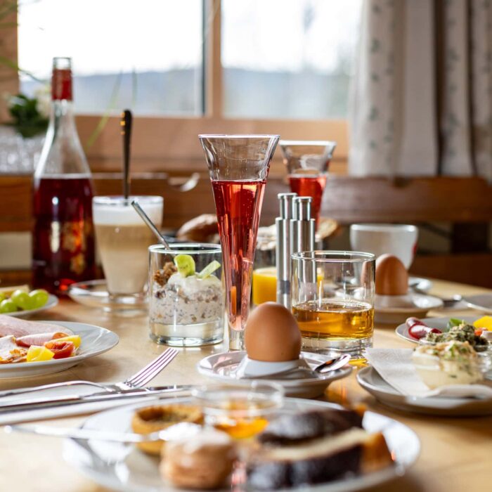 Frühstückstisch mit einer Vielzahl von Speisen und Getränken, darunter frisches Obst, Wurst, Käse, Joghurt, ein gekochtes Ei, verschiedene Getränke wie Saft und Kaffee sowie ein Glas mit Müsli. Der Tisch ist hell dekoriert und bietet eine einladende Atmosphäre für ein reichhaltiges Frühstück.