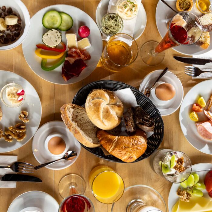 Vielfältiges Frühstücksbuffet auf Holztisch mit verschiedenen Speisen wie frischem Brot, Aufschnitt, Käse, Obst, Joghurt, Gemüse und Getränken wie Orangensaft und Bier. Ideal für einen energiereichen Start in den Tag.