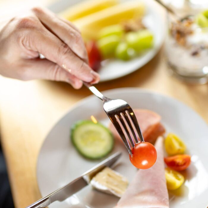 Eine Hand hält eine Gabel mit einer Kirschtomate, während auf einem Teller verschiedene Lebensmittel wie Gurken, Schinken und Obst zu sehen sind. Im Hintergrund befindet sich ein weiteres Gericht, das Joghurt und Früchte enthält. Das Bild vermittelt eine einladende Frühstücksszene.