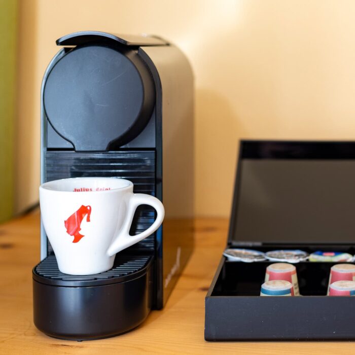 Nespresso Maschine mit einer Tasse Kaffee und einer Box mit Kaffeekapseln auf einem Holztisch. Die Tasse trägt ein rotes Logo und steht auf der Abtropfschale der Maschine. Im Hintergrund ist eine Steckdose sichtbar.