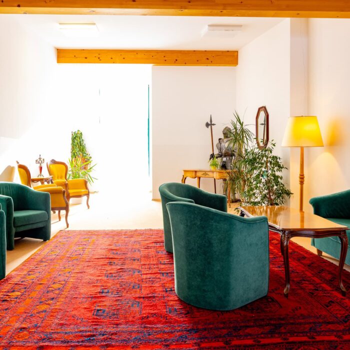 Helles Wohnzimmer mit eleganten grünen Sesseln und einer roten Teppichgestaltung. Im Hintergrund stehen gelbe Stühle und ein Holztisch. Pflanzen und eine Stehlampe ergänzen die gemütliche Atmosphäre. Ideal für Inneneinrichtungsinspiration.