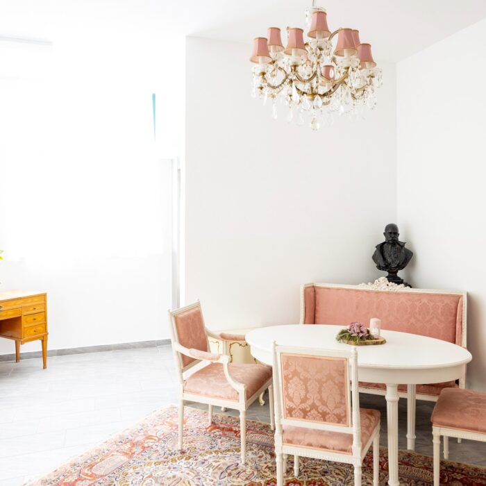 Heller, einladender Essbereich mit ovalem Tisch und eleganten Stühlen in zartem Rosa, ergänzt durch einen antiken Schreibtisch aus Holz und eine dekorative Pflanze. Im Hintergrund befindet sich eine stilvolle Deckenleuchte und eine Büste, die dem Raum einen nostalgischen Charme verleiht.