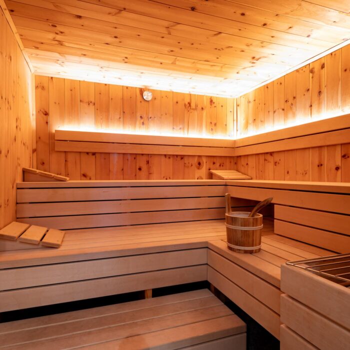 Sauna mit hellen Holzverkleidungen, Bänken und einem Holzbehälter für Wasser, ideal für Entspannung und Wellness. Die sanfte Beleuchtung schafft eine angenehme Atmosphäre.