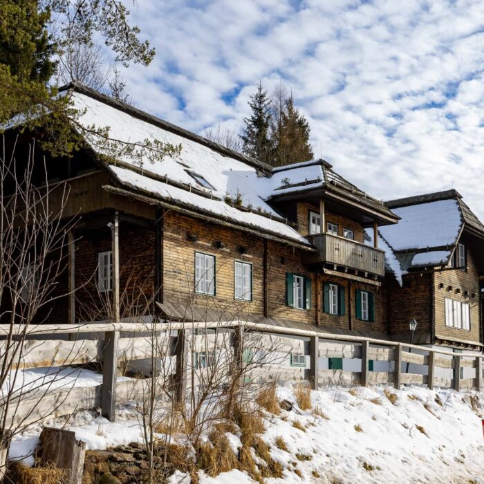 Historische Holzhäuser in einer winterlichen Landschaft mit Schnee und bewaldetem Hintergrund, ideal für Naturliebhaber und Architekturinteressierte. Die Gebäude sind mit einem traditionellen Stil und grünen Fensterläden gestaltet, während der Himmel mit Wolken durchzogen ist.