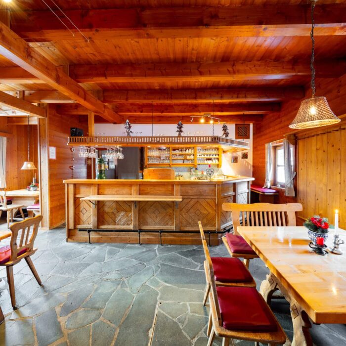 Gemütlicher Essbereich in einer rustikalen Holzhausküche mit einem großen Tisch, Stühlen mit roten Polstern und einer einladenden Bar. Der Raum ist mit warmen Holzvertäfelungen und einem eleganten Steinboden gestaltet, ideal für gesellige Zusammenkünfte.