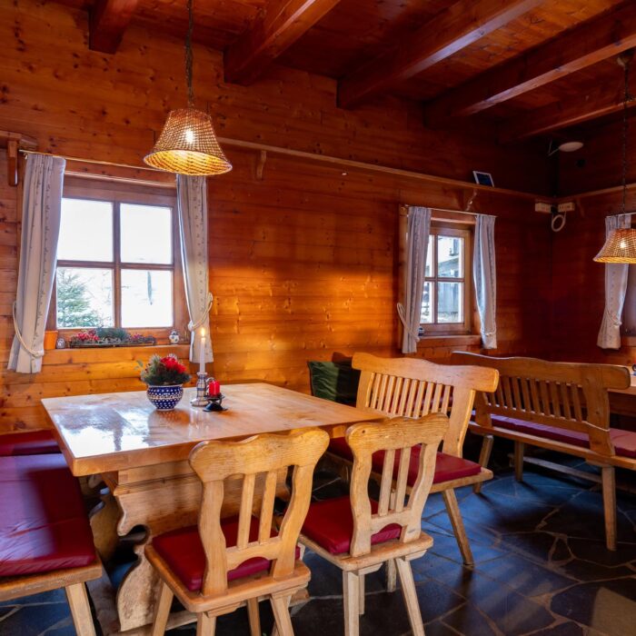 Gemütlicher Gastraum in einem Holzhaus mit einem großen Holztisch und Bänken. Die warmen Holztöne und die sanfte Beleuchtung schaffen eine einladende Atmosphäre. Ideal für gesellige Runden oder entspannte Abende.
