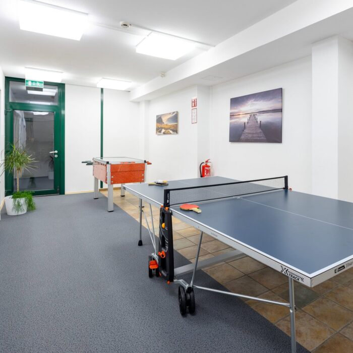 Tischtennisraum mit einem Tischtennistisch, einer roten Tischtennisplatte und einem modernen, hellen Ambiente. An den Wänden hängen Bilder, während der Boden mit Teppich ausgelegt ist. Ideal für Freizeitaktivitäten und sportliche Betätigung.