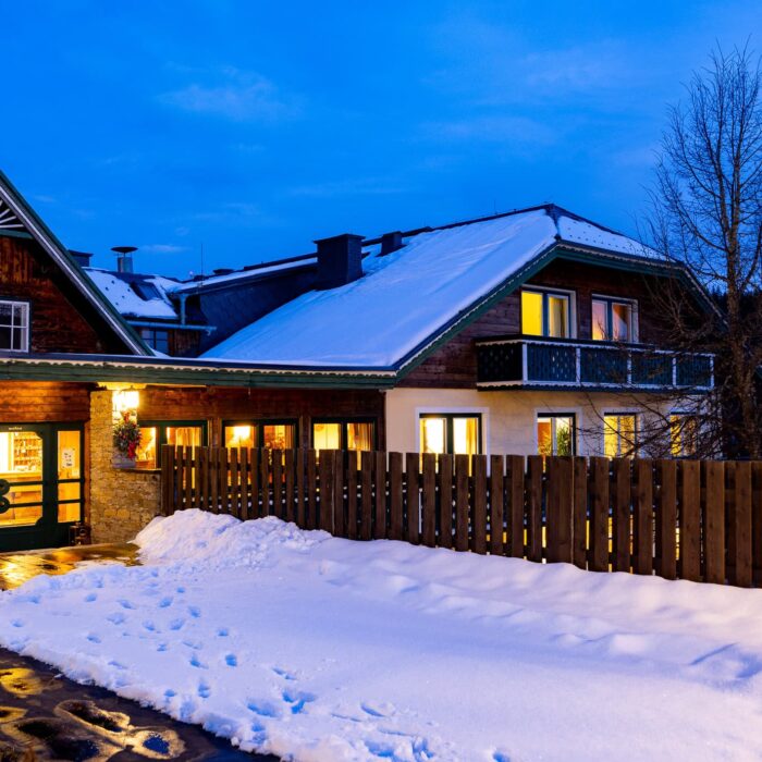 Das Bild zeigt ein charmantes, winterliches Gebäude mit einem Holz- und Steinmix, das in der Dämmerung beleuchtet ist. Der schneebedeckte Eingangsbereich und die Holzlattenzaun verleihen eine einladende Atmosphäre. Ideal für eine Unterkunft in der Natur oder für einen Winterurlaub.