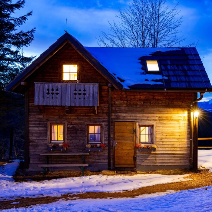 Gemütliches Holzhaus im Schnee bei Dämmerung, umgeben von Bäumen und Bergen. Die Fenster sind beleuchtet, was eine einladende Atmosphäre schafft. Ideal für Winterurlaube oder romantische Ausflüge in die Natur.