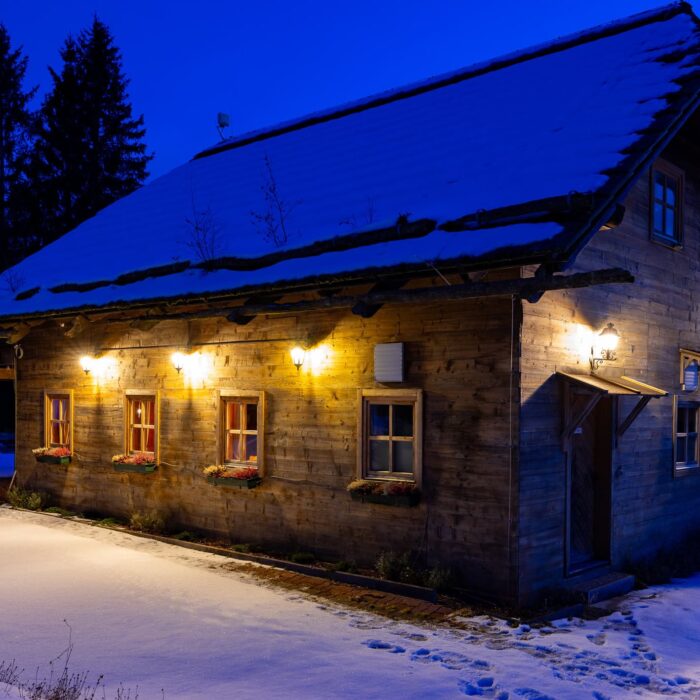 Gemütliches Holzhaus in der Dämmerung, umgeben von Schnee, mit warmem Licht an den Fenstern und einer einladenden Atmosphäre. Die Landschaft ist ruhig und der Himmel hat einen tiefblauen Farbton, der die winterliche Szene unterstreicht. Ideal für Winterurlaube oder romantische Ausflüge.