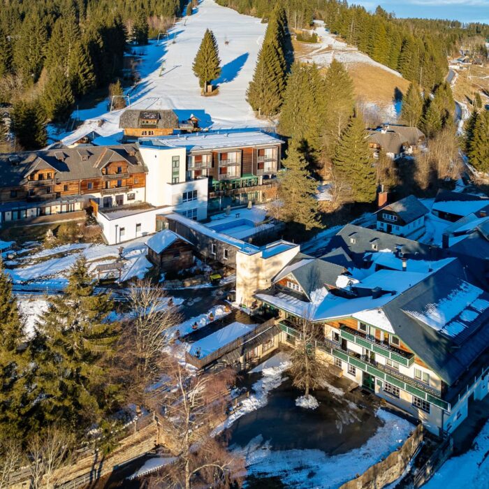 Luftaufnahme eines winterlichen Ferienortes mit mehreren Gebäuden, umgeben von schneebedeckten Landschaften und Tannenbäumen. Zu sehen sind traditionelle und moderne Architekturelemente, die eine einladende Atmosphäre schaffen. Ideal für Winterurlaub und Erholung in der Natur.