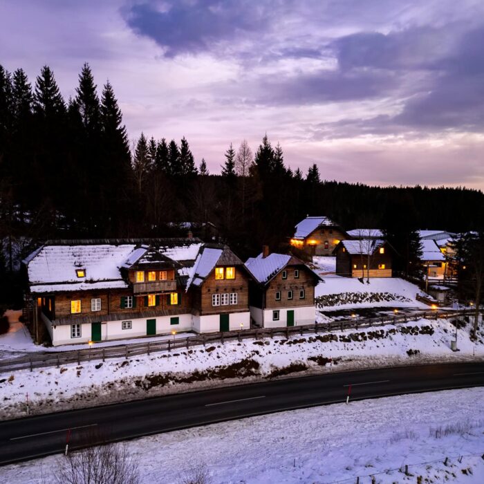 Schneebedeckte Landschaft mit traditionellen Holzchalets im Abendlicht, umgeben von einem Wald. Die Gebäude sind beleuchtet und spiegeln eine gemütliche Atmosphäre wider, während die Straße im Vordergrund sanft durch die Winterlandschaft führt.