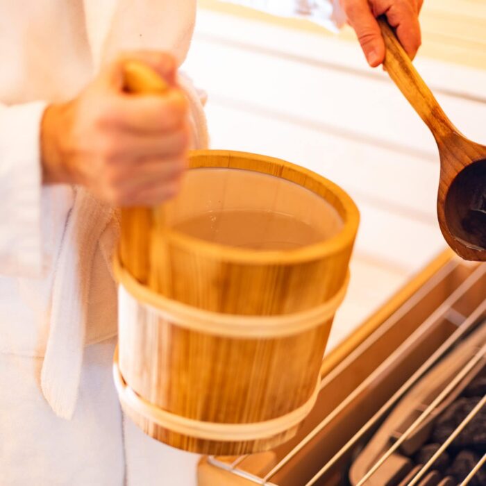 Person hält einen Holzbehälter in der Hand, während sie mit einem Holzlöffel in einer Sauna oder einem Wellnessbereich arbeiten. Der Fokus liegt auf der natürlichen Holzoptik des Behälters und der entspannten Atmosphäre.
