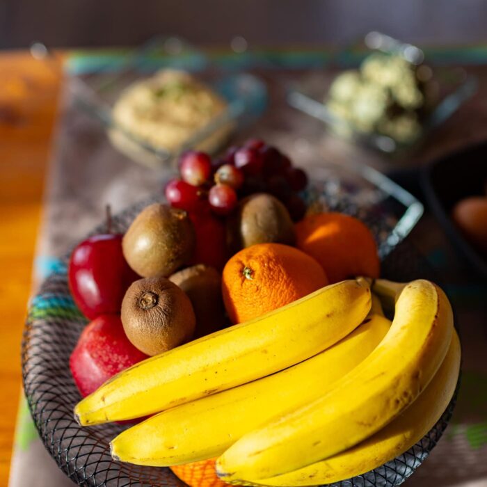 Bunte Obstschale mit Bananen, Äpfeln, Trauben und Kiwis, arrangiert auf einem Tisch, im Hintergrund weitere Speisen.