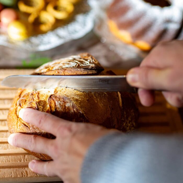 Ein Mensch schneidet frisch gebackenes Brot mit einem scharfen Messer auf einem Holzbrett, während im Hintergrund verschiedene süße Leckereien wie Macarons und Gebäck zu sehen sind.