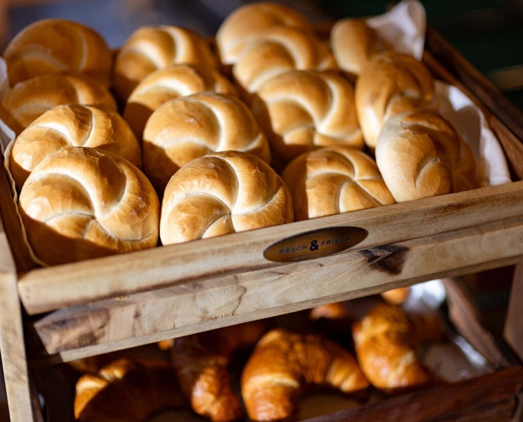 Frisch gebackene Brötchen in einem Holzkorb, darunter Croissants, ideal für ein Frühstücksbuffet oder eine Bäckerei. Die Brötchen haben eine goldbraune Kruste und sind sorgfältig angerichtet.
