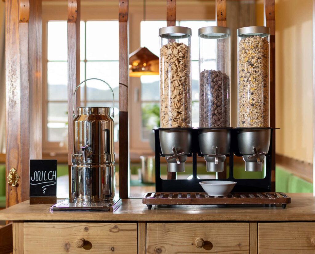 Müsli- und Milchstation in einem gemütlichen Ambiente, mit dekorativen Glasbehältern für verschiedene Müslisorten und einem silbernen Milchspender auf einem Holzregal. Ideal für ein Frühstücksbuffet oder eine gesunde Snackauswahl.