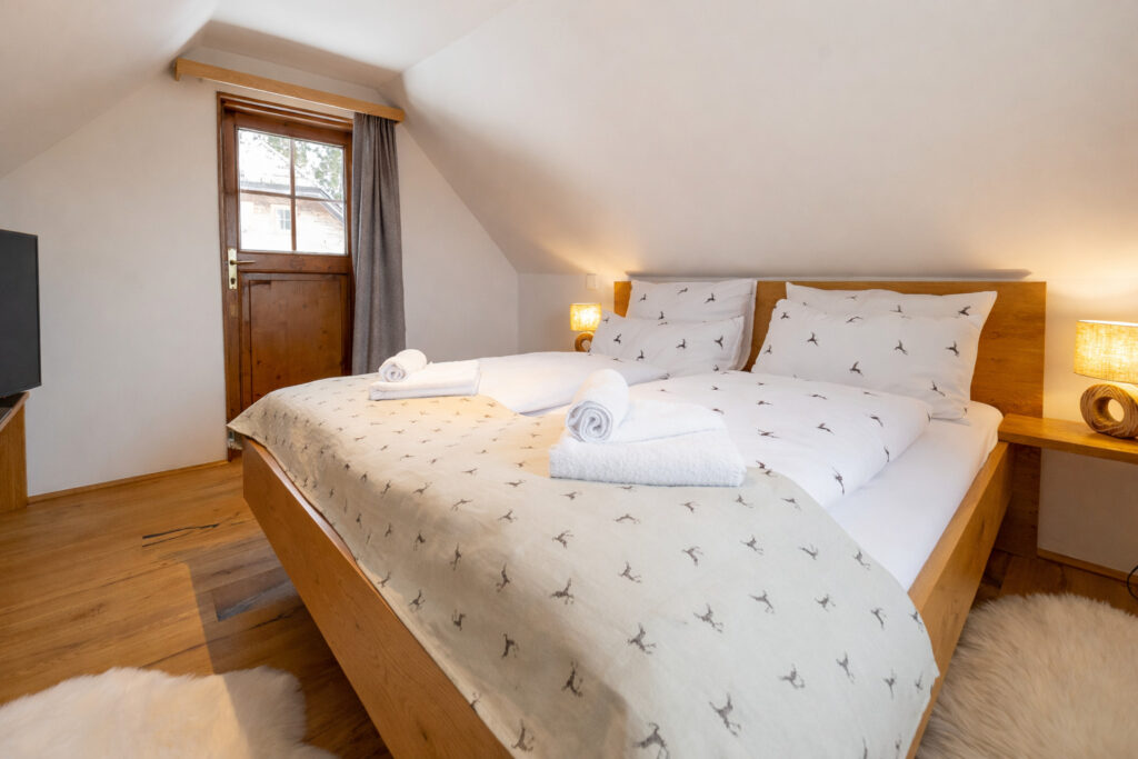 Gemütliches Schlafzimmer mit Doppelbett, weißen Bettwäsche und handtuch, Holzdetails und einem Fenster, das natürliches Licht hereinlässt. Der Raum verfügt über einen Fernseher und eine warme Atmosphäre, ideal für erholsame Nächte.