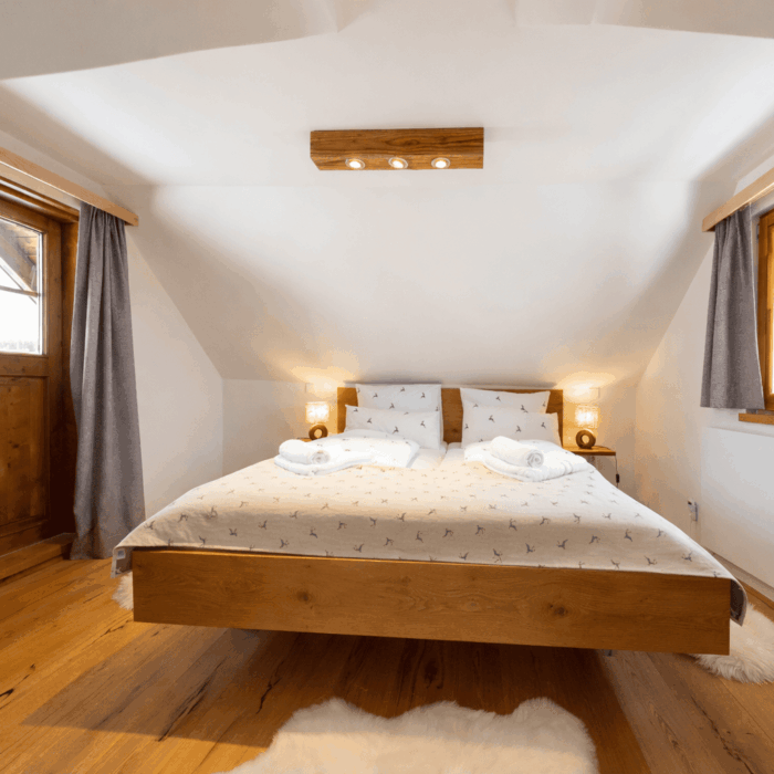 Gemütliches Schlafzimmer mit Doppelbett, Holzdetails und sanfter Beleuchtung, ideal für einen erholsamen Aufenthalt in den Bergen. Helle Wände und Fenster mit Vorhängen sorgen für eine einladende Atmosphäre.