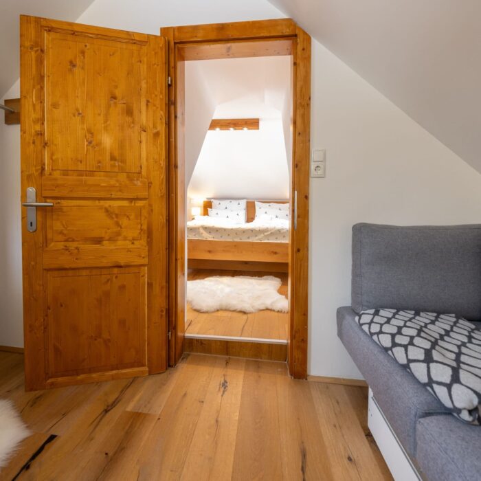 Gemütlicher Raum mit Holzdetails, der durch eine offene Tür zu einem Schlafzimmer führt. Sichtbare Möbel umfassen eine graue Couch und einen Holzfußboden. Im Hintergrund ist ein Bett mit Kissen und einer Decke zu erkennen, was eine einladende Atmosphäre schafft. Ideal für eine Ferienunterkunft oder ein modernes Zuhause.