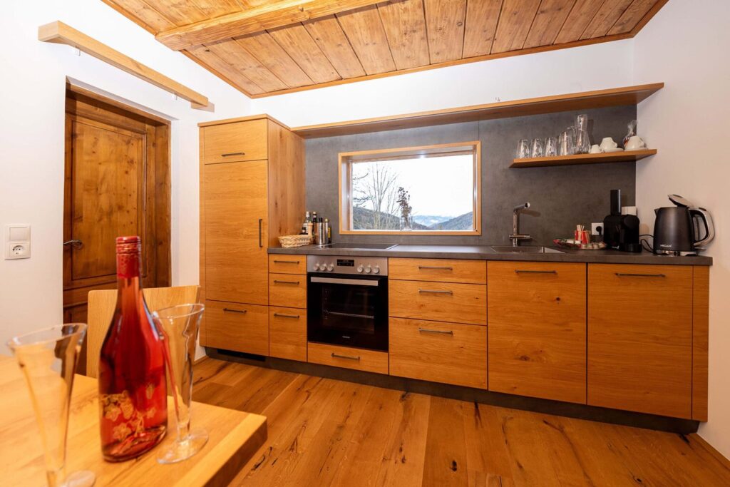 Moderne Küche mit Holzschränken und Edelstahlgeräten, ausgestattet mit einem Herd, einem Wasserkocher und Gläsern. Im Vordergrund steht eine Flasche Roséwein auf einem Holztisch, während ein Fenster einen Blick auf die umliegende Natur bietet.