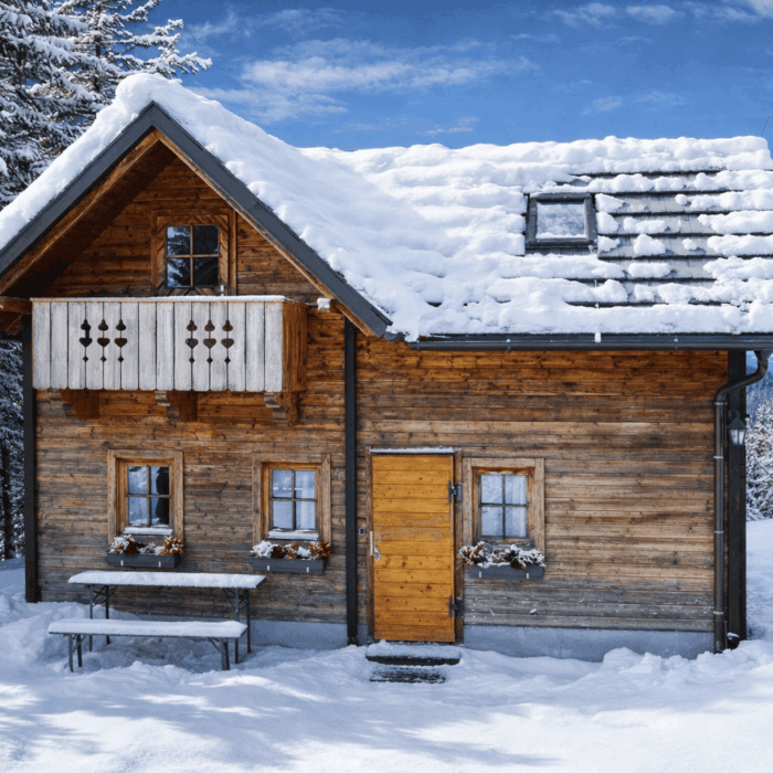 Gemütliches Holzhaus im Winter mit schneebedecktem Dach und Balkon, umgeben von verschneiten Bäumen und klarem blauen Himmel. Ideal für eine entspannte Auszeit in der Natur.