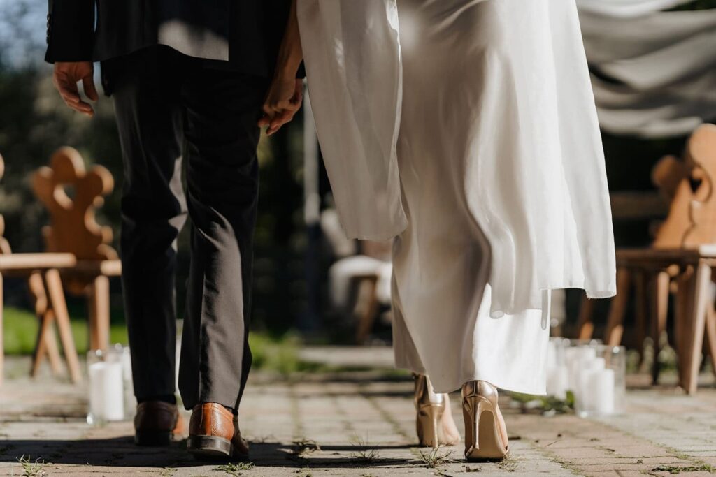 Hochzeitspaar, das Hand in Hand einen Weg entlanggeht, während sie in eleganter Hochzeitskleidung gekleidet sind. Im Hintergrund sind stilvolle Holzstühle und Kerzen zu sehen, die eine romantische Atmosphäre schaffen.