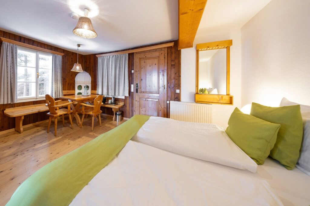 Gemütliches Zimmer mit zwei Einzelbetten und grünen Kissen, Holzdecke und -wänden, einem Tisch mit Stühlen sowie einem Fenster, das Tageslicht hereinlässt. Ideal für einen entspannten Aufenthalt in den Bergen.