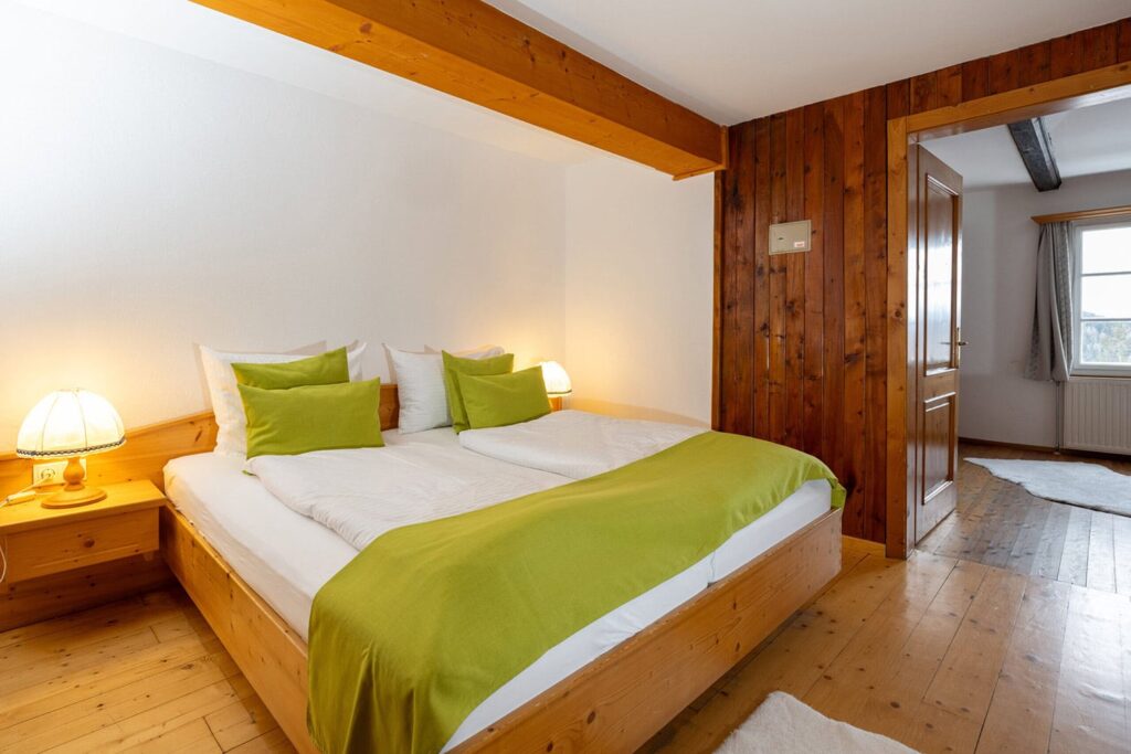 Gemütliches Schlafzimmer mit einem Doppelbett, ausgestattet mit grünen Kissen und einer weißen Bettdecke. Die warmen Holzelemente und die sanfte Beleuchtung schaffen eine einladende Atmosphäre. Im Hintergrund ist eine Tür zu einem weiteren Raum sichtbar, während ein Fenster Tageslicht hereinlässt. Ideal für einen erholsamen Aufenthalt.