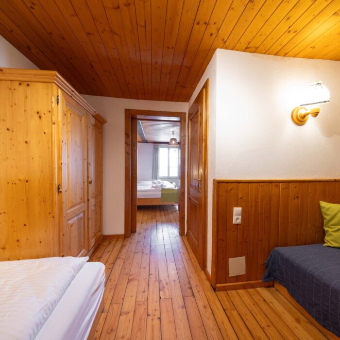 Gemütlicher Innenraum eines Holzhauses mit Sicht auf einen Flur, der zu einem weiteren Raum führt. Die Wände sind mit Holz verkleidet, und es gibt einen Schlafbereich mit einem Sofa und einem Kleiderschrank. Helle Beleuchtung sorgt für eine einladende Atmosphäre.