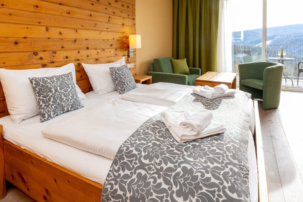 Gemütliches Hotelzimmer mit zwei Betten, dekoriert mit grauen Kissen und einem schönen Überwurf. Im Hintergrund sind ein grüner Sessel und ein Tisch zu sehen, während große Fenster den Raum mit natürlichem Licht fluten und einen Blick auf die umliegende Landschaft bieten. Frische Handtücher sind ordentlich auf dem Bett platziert.