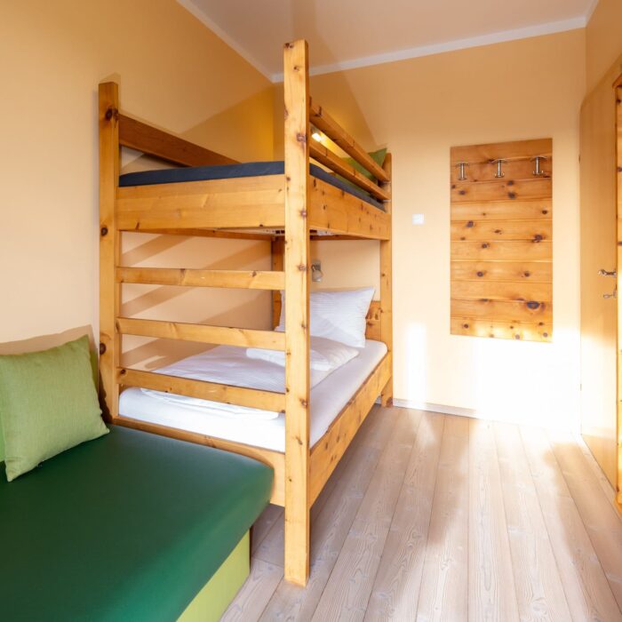 Gemütliches Zimmer mit Etagenbett aus Holz, einem grünen Sofa und natürlichem Licht. Ideal für Familien oder Gruppen, die eine komfortable Unterkunft suchen. Helle Wände und Holzdetails schaffen eine einladende Atmosphäre.