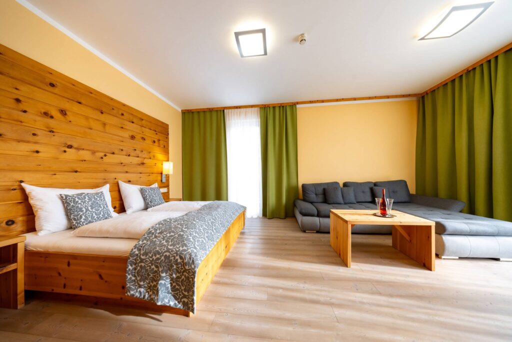 Helles und modernes Zimmer mit Holzverkleidung, Doppelbett mit dekorativen Kissen, einem gemütlichen grauen Sofa und einem Holzkaffee-Tisch. Große Fenster mit grünen Vorhängen sorgen für viel Tageslicht und eine einladende Atmosphäre. Ideal für entspannende Aufenthalte.