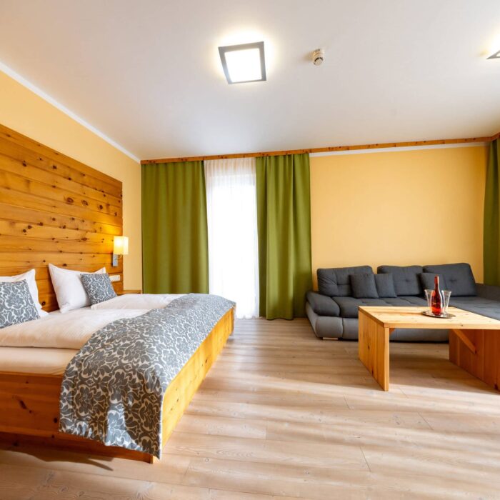 Helles und modernes Zimmer mit Holzverkleidung, Doppelbett mit dekorativen Kissen, einem gemütlichen grauen Sofa und einem Holzkaffee-Tisch. Große Fenster mit grünen Vorhängen sorgen für viel Tageslicht und eine einladende Atmosphäre. Ideal für entspannende Aufenthalte.