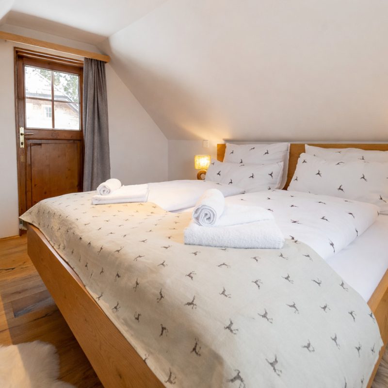 Gemütliches Schlafzimmer mit Doppelbett, weißen Bettwäsche und handtuch, Holzdetails und einem Fenster, das natürliches Licht hereinlässt. Der Raum verfügt über einen Fernseher und eine warme Atmosphäre, ideal für erholsame Nächte.