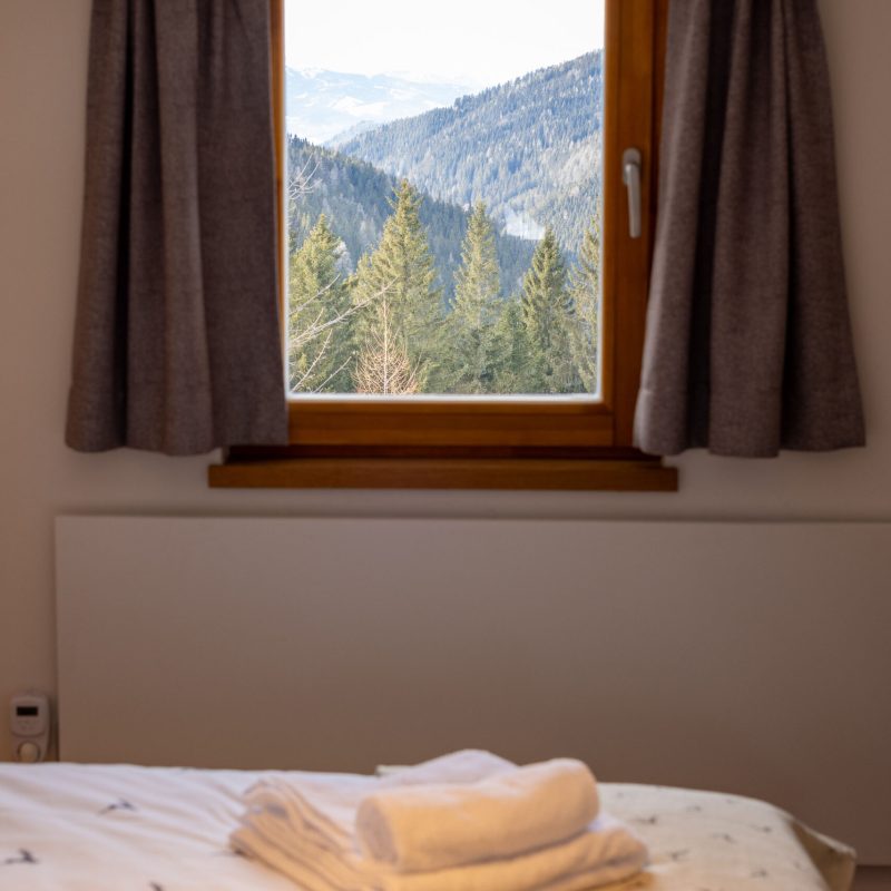 Blick aus einem Schlafzimmerfenster auf eine beeindruckende Berglandschaft mit Nadelbäumen und Hügeln, im Vordergrund ein Bett mit Kissen und Handtüchern, das eine gemütliche Atmosphäre vermittelt.