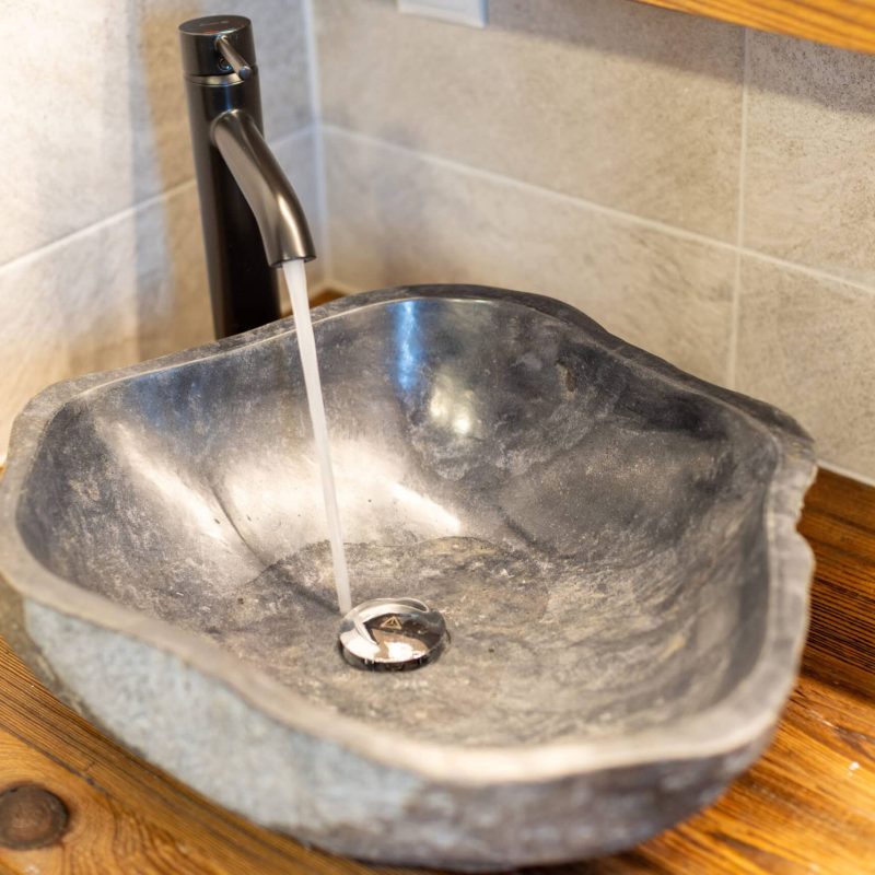 Natursteinwaschbecken aus schwarzem Marmor auf einem Holzuntergrund, mit einem modernen, schwarzen Wasserhahn, der klaren Wasserstrahl abgibt. Ideal für stilvolle Badezimmergestaltungen.