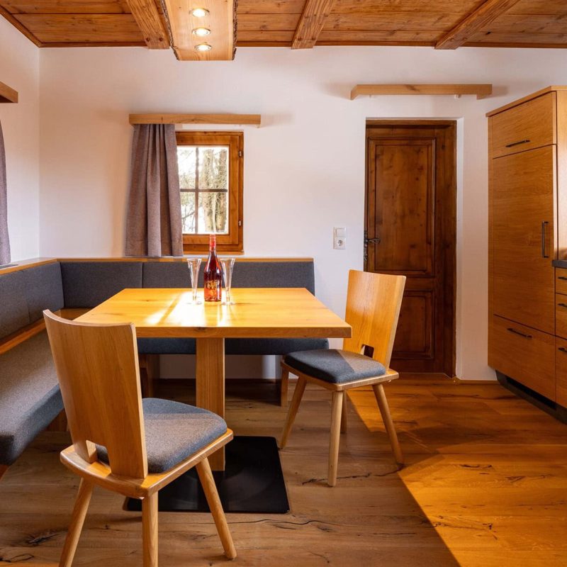 Moderne Küche mit Holzvertäfelung, einem Essbereich mit grauem Sitzpolster, einem Holztisch und Stühlen. Die Küche ist ausgestattet mit einem Herd, Ofen und Küchenutensilien. Große Fenster sorgen für viel Tageslicht. Ideal für gemütliche Mahlzeiten oder gesellige Runden.