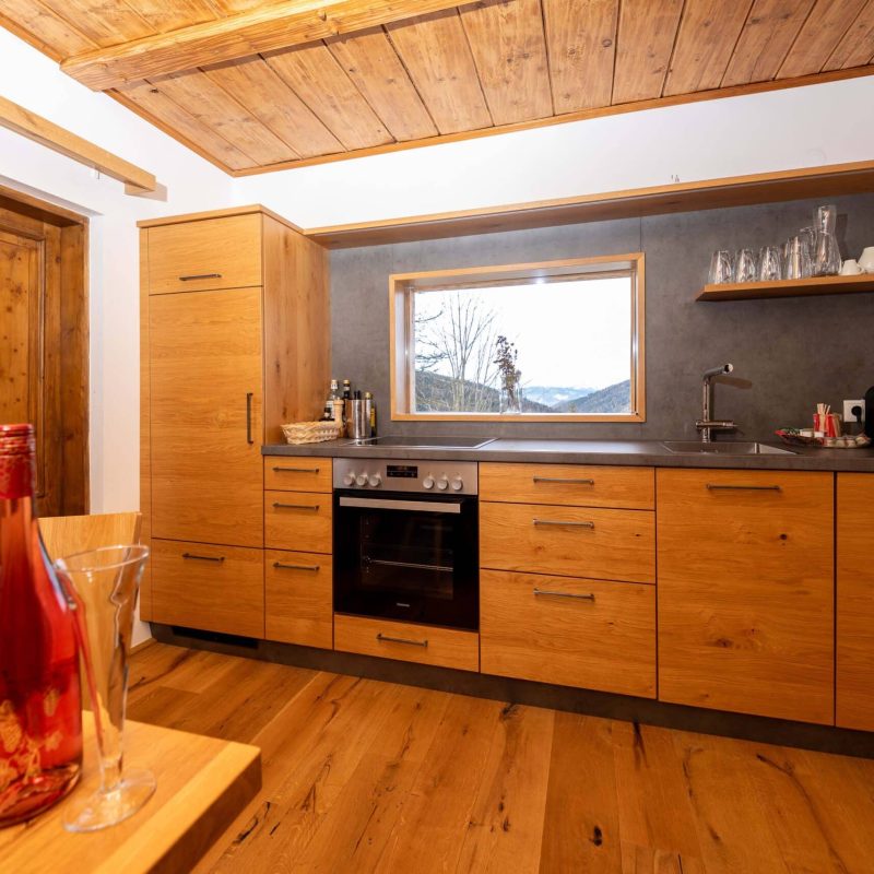 Moderne Küche mit Holzmöbeln, ausgestattet mit einem Herd und einem Fenster, das einen Blick auf die Umgebung bietet. Der Boden ist aus Holz, und im Vordergrund stehen Gläser und eine Flasche.