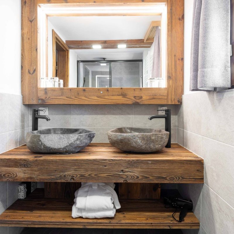 Modernes Badezimmer mit zwei eleganten Waschbecken aus Stein auf einem rustikalen Holztisch, beleuchtet von einem großen Spiegel. Die Wände sind mit grauen Fliesen verkleidet, und im Hintergrund ist eine Duschkabine sichtbar. Handtücher liegen ordentlich gestapelt auf einem offenen Regal.